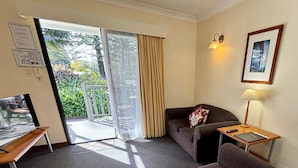 Une chambre d’hôtel avec un canapé, une petite table avec une lampe, un téléviseur posé sur un meuble, une porte-fenêtre coulissante donnant sur un balcon et un tableau encadré au mur.