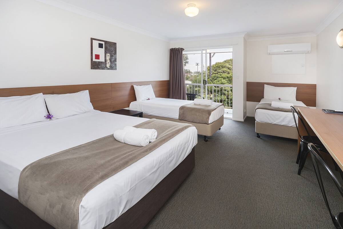 A hotel room with two beds, a desk, and a balcony with a view.