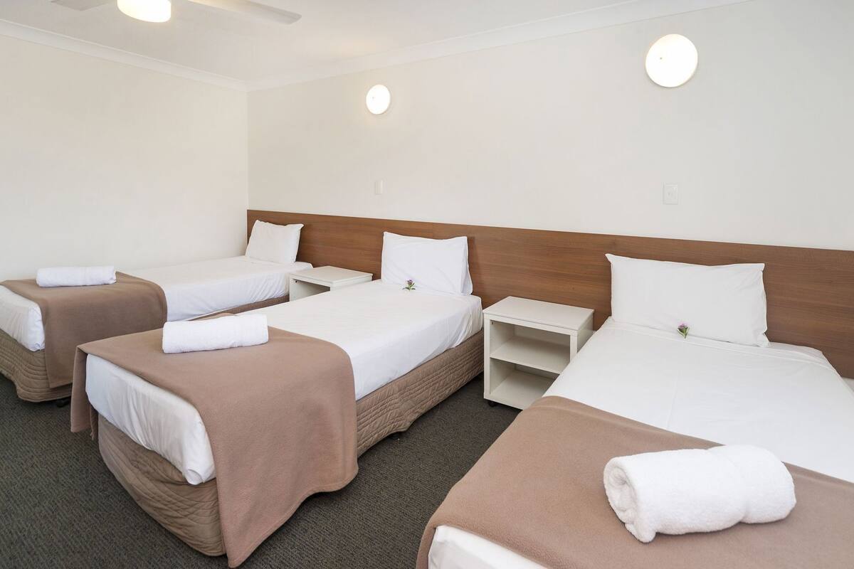A hotel room with three beds, each with a pillow and a rolled towel. There are bedside tables and a wall-mounted light fixture.