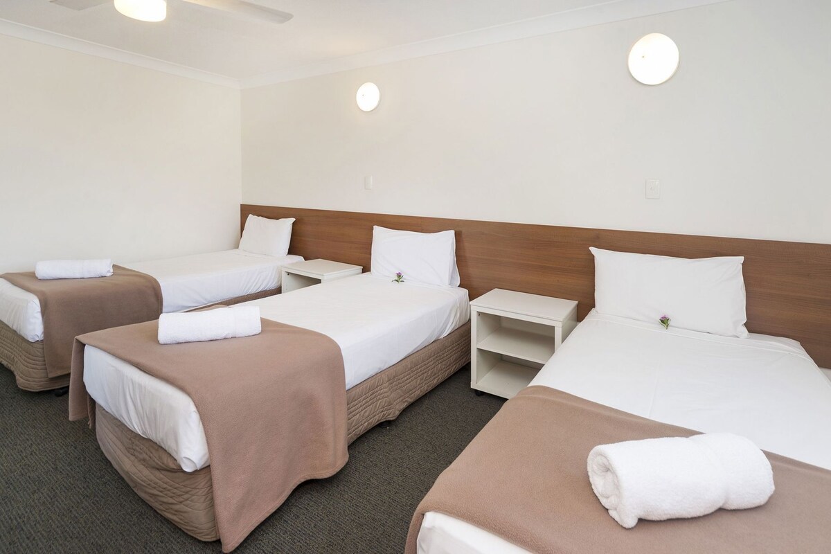 Habitación de hotel con tres camas, cada una con almohada y una toalla enrollada. Hay mesitas de noche y una lámpara de pared.
