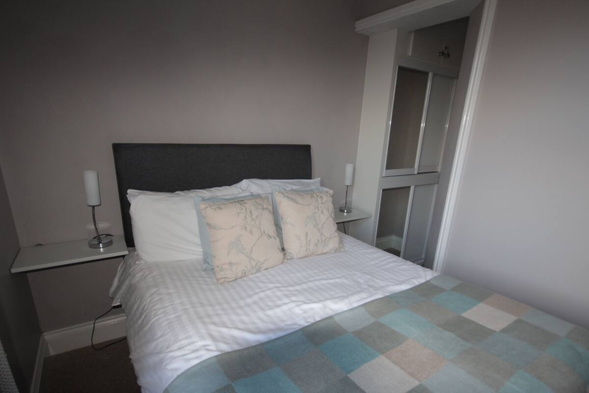 A neatly made bed with a checkered bedspread, two bedside tables with lamps, and a door leading to another room.