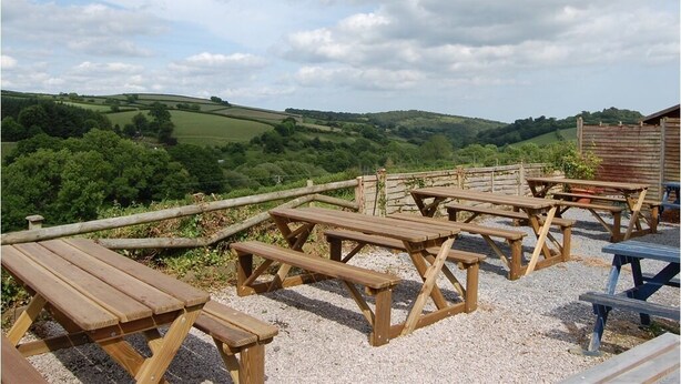 Outdoor wooden picnic tables arranged on a gravel surface with a view of rolling hills and a clear sky. - The Tradesmans Arms (Buckfastleigh)
