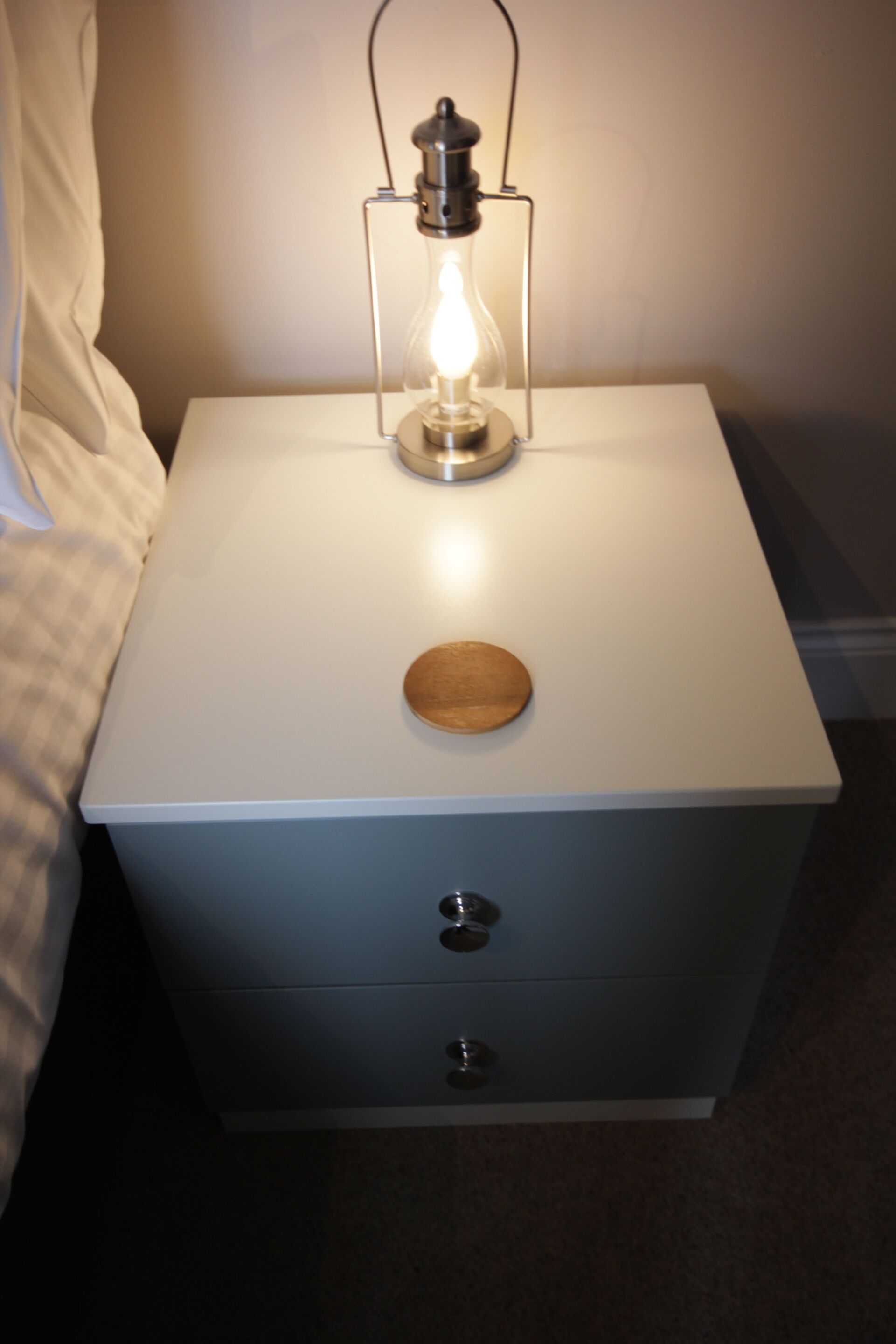 A white bedside table with a lit Edison bulb lamp, a wooden knob, and two drawers.