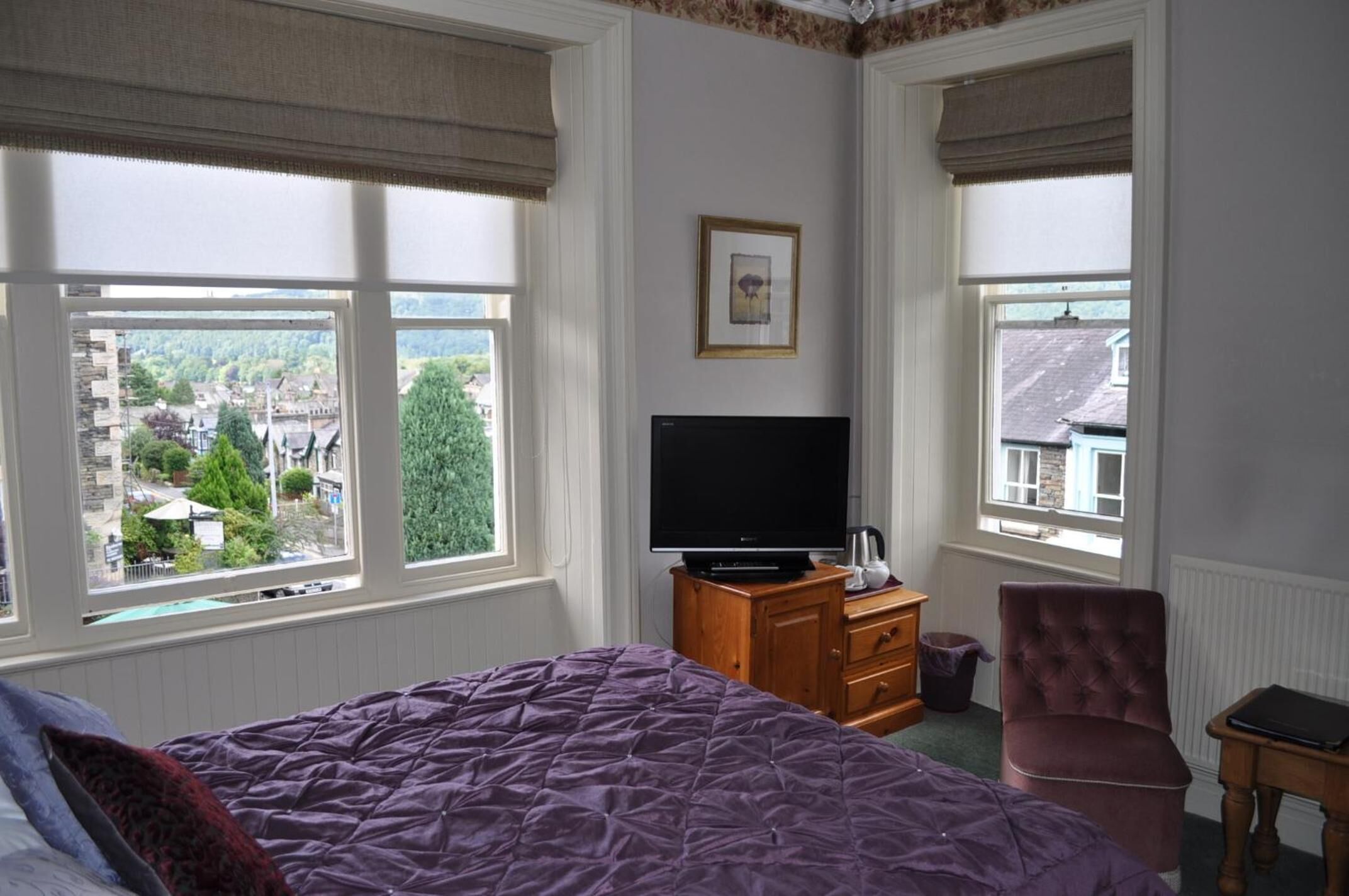 A bedroom with a bed, a television on a wooden dresser, a chair, and large windows with views of a town.