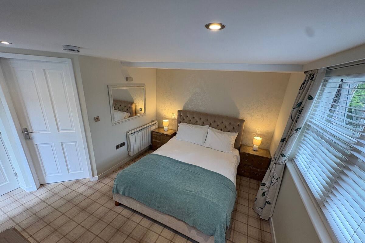 A bedroom with a bed, bedside lamps, a mirror, a radiator, and a window with blinds.