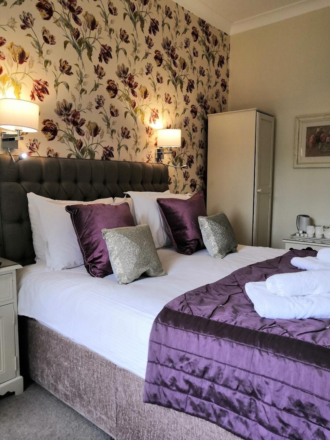 A neatly made bed with a purple and white duvet, a dark upholstered headboard, and decorative pillows.