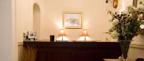 A reception area with a wooden reception desk, two lamps, a chandelier, a framed picture, and a vase with flowers.