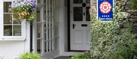 Un bâtiment en pierre avec une porte blanche, des jardinières suspendues et une enseigne indiquant qu’il s’agit d’une maison d’hôtes.