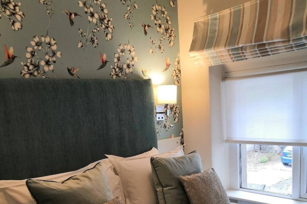 A neatly made bed with pillows and a folded towel, a window with a striped blind, and a wall with floral wallpaper.
