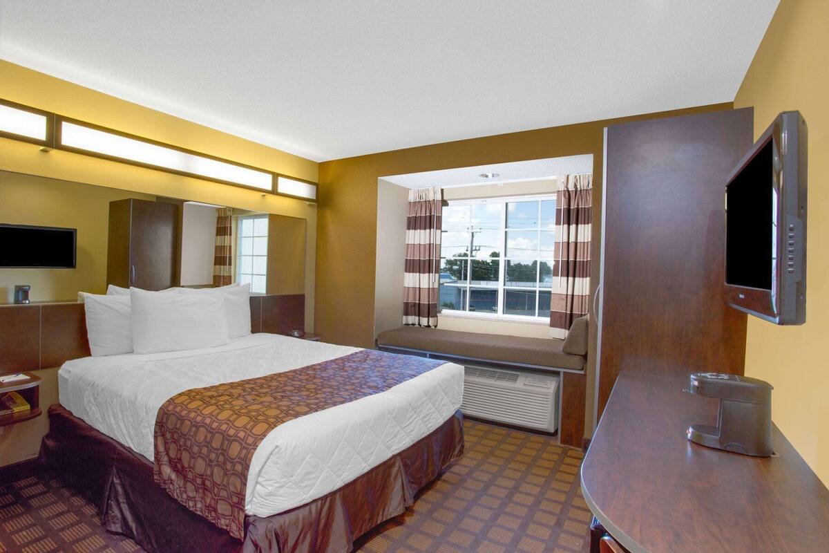 A hotel room with a bed, a television, a window with curtains, a desk, and a heater.