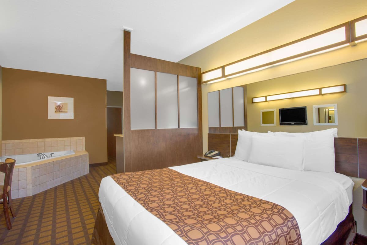 A hotel room with a large bed, a separate bathroom with a bathtub, a desk with a chair, and a flat-screen TV mounted on the wall.