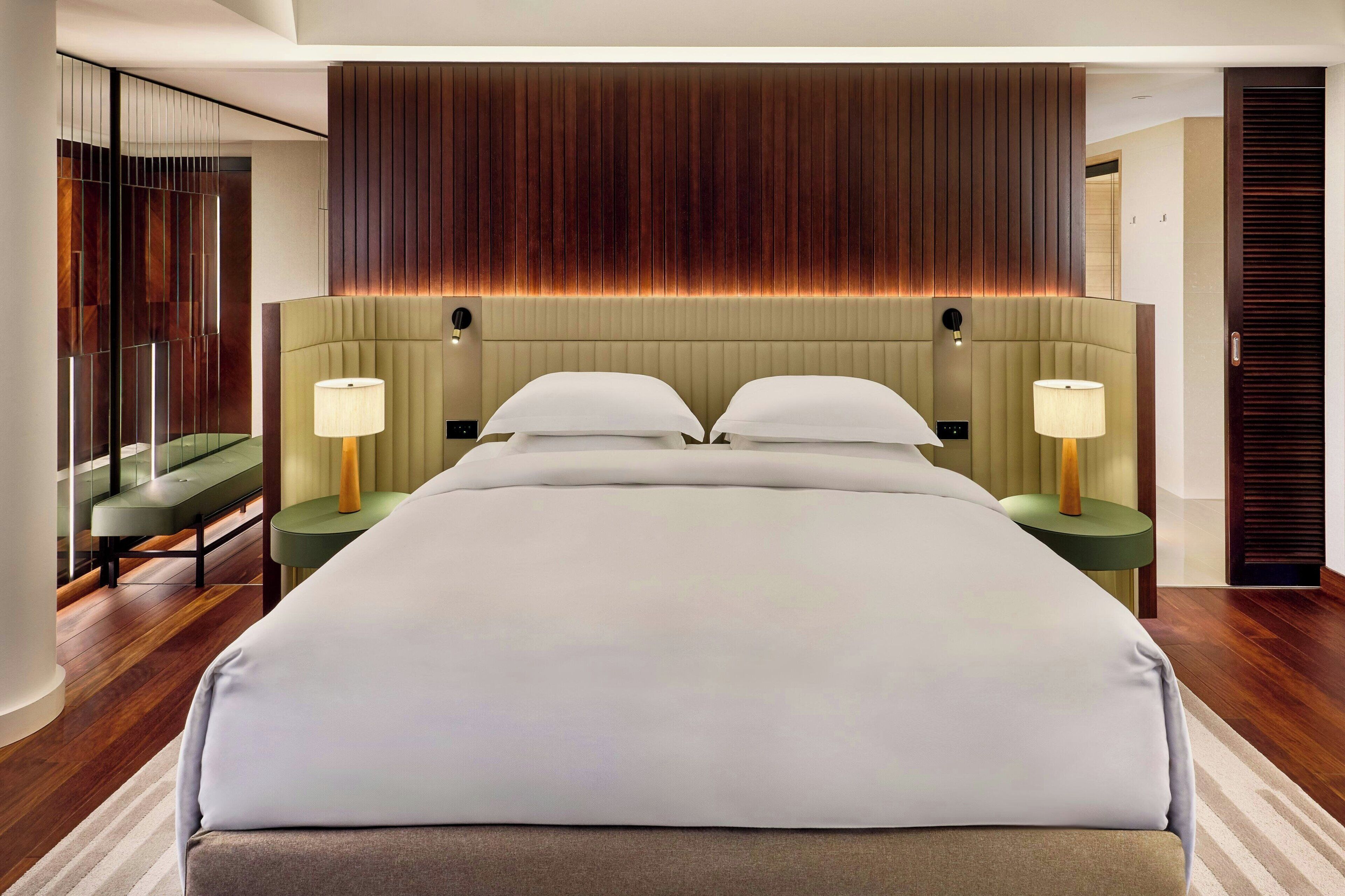 Una habitación de hotel moderna con una cama grande, dos mesitas de noche con lámparas, un banco verde y un panel de pared de madera.