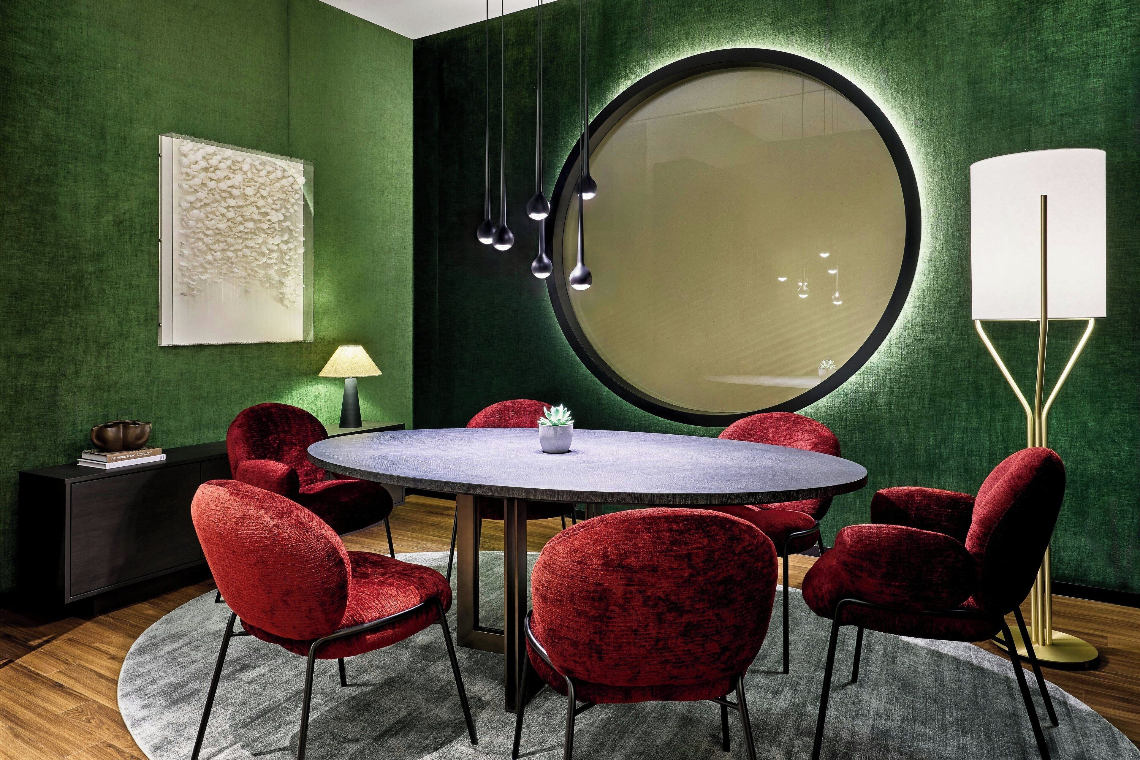 Una sala conferenze con un tavolo rotondo, sedie rosse, un grande specchio e una lampada da terra.