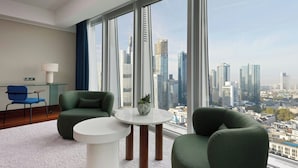 A modern hotel room with a large window offering a city view, a round white table, two green armchairs, and a blue chair.