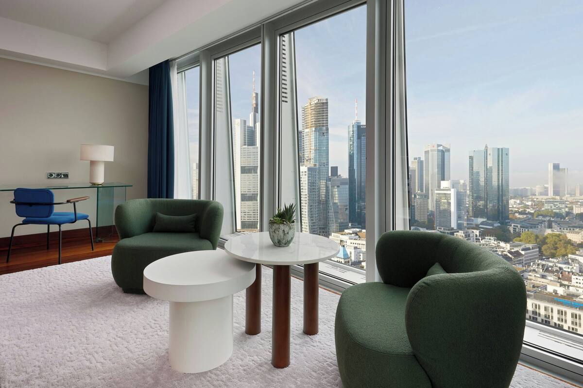 A modern hotel room with a large window offering a city view, a round white table, two green armchairs, and a blue chair.