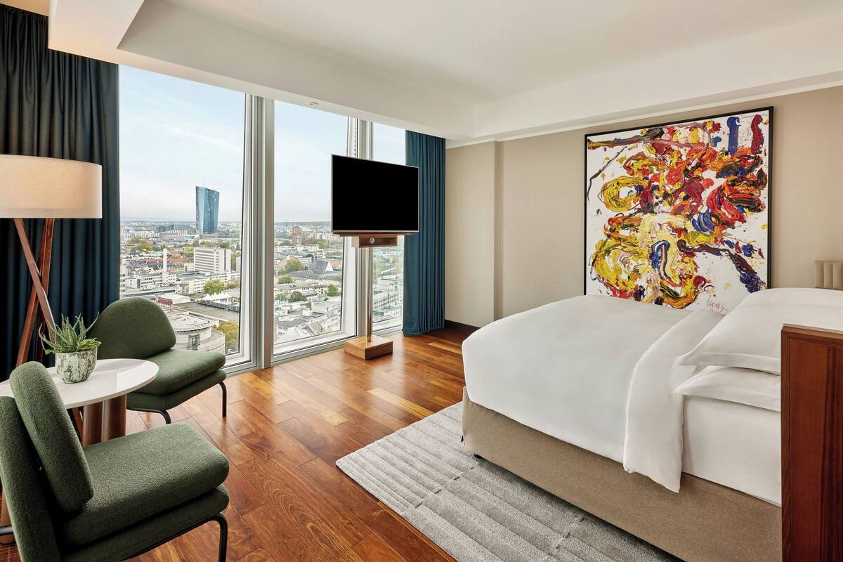 A hotel room with a large bed, a TV, a desk with a chair, and a view of the city.