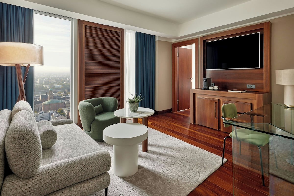 A modern hotel room with a large window, a sofa, a glass table, a chair, a small round table, and a TV.