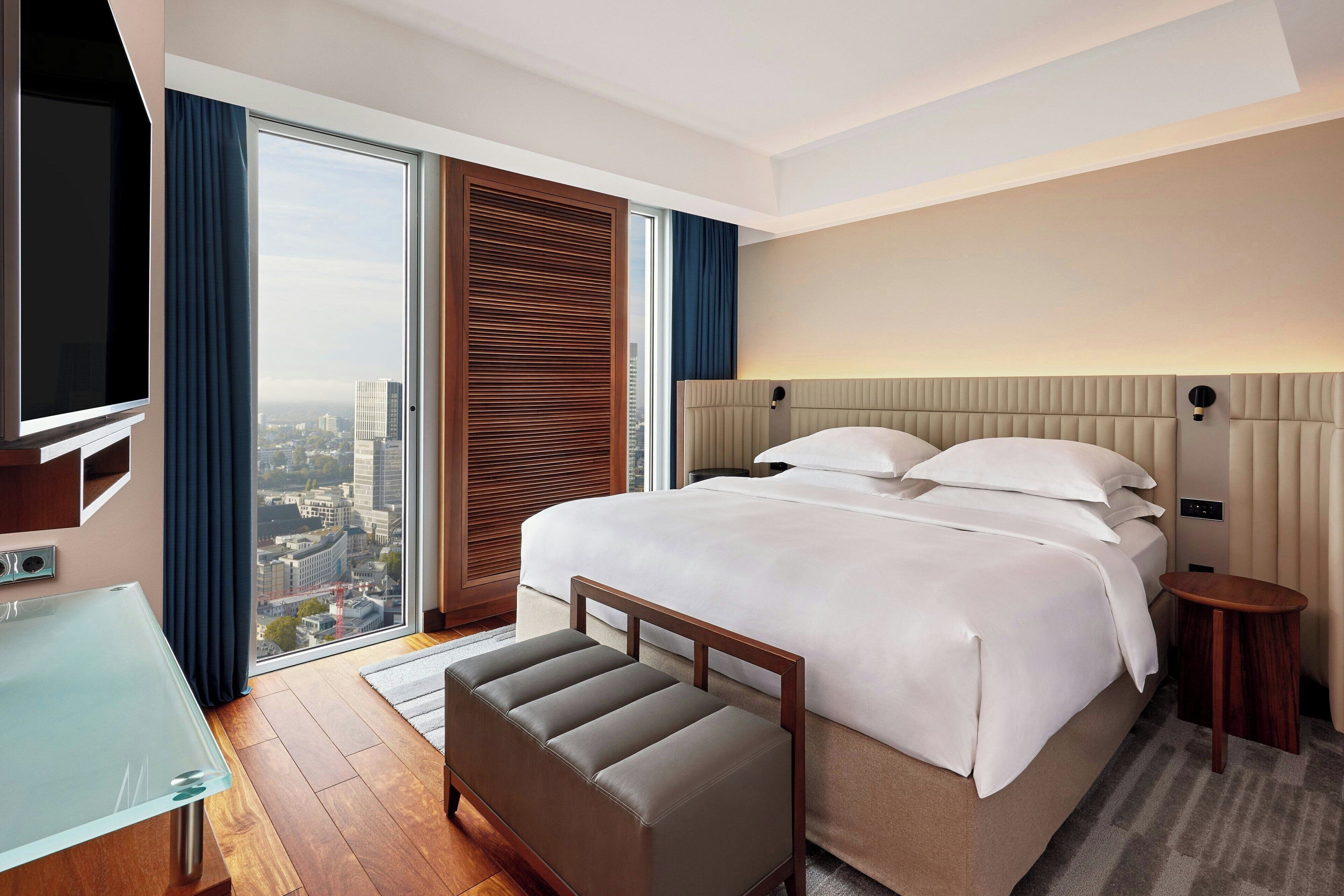 A modern hotel room with a large bed, a glass TV, and a view of the city.