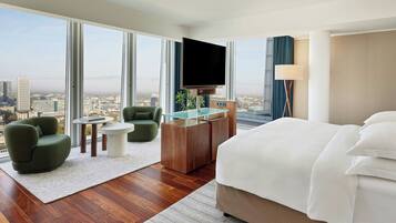 A hotel room with a large bed, a desk with a flat-screen TV, a green armchair, and a view of the city.