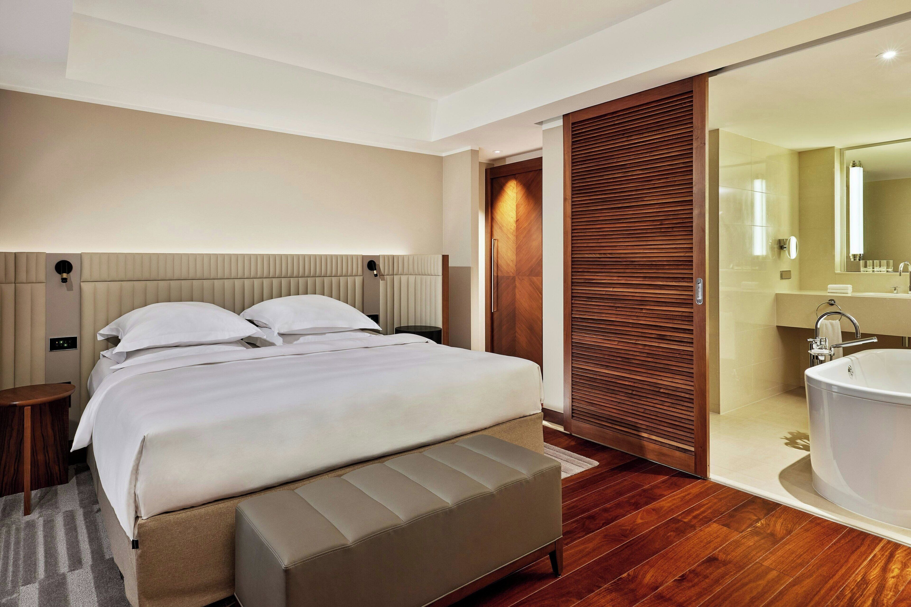 Una habitación de hotel moderna con una cama grande, un panel de pared de madera, un baño con bañera y un espejo amplio.