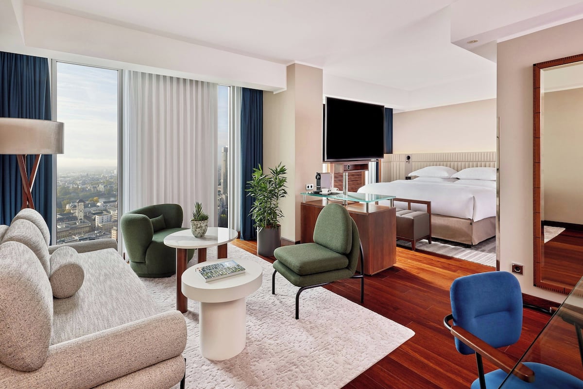 A modern hotel room with a large bed, a flat-screen TV, a sofa, a chair, a small table, and a potted plant.
