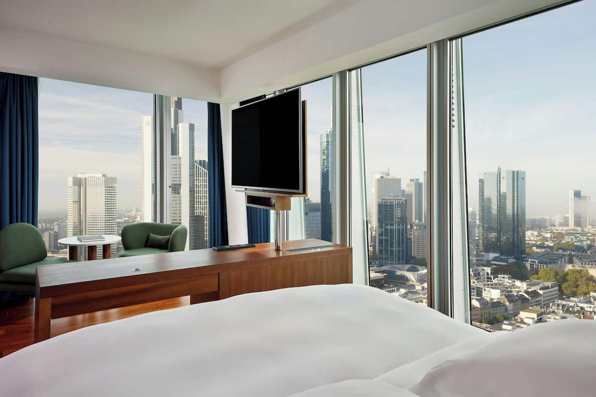 A hotel room with a large bed, a TV on a wooden stand, a small table, two green chairs, and a view of a cityscape through large windows.
