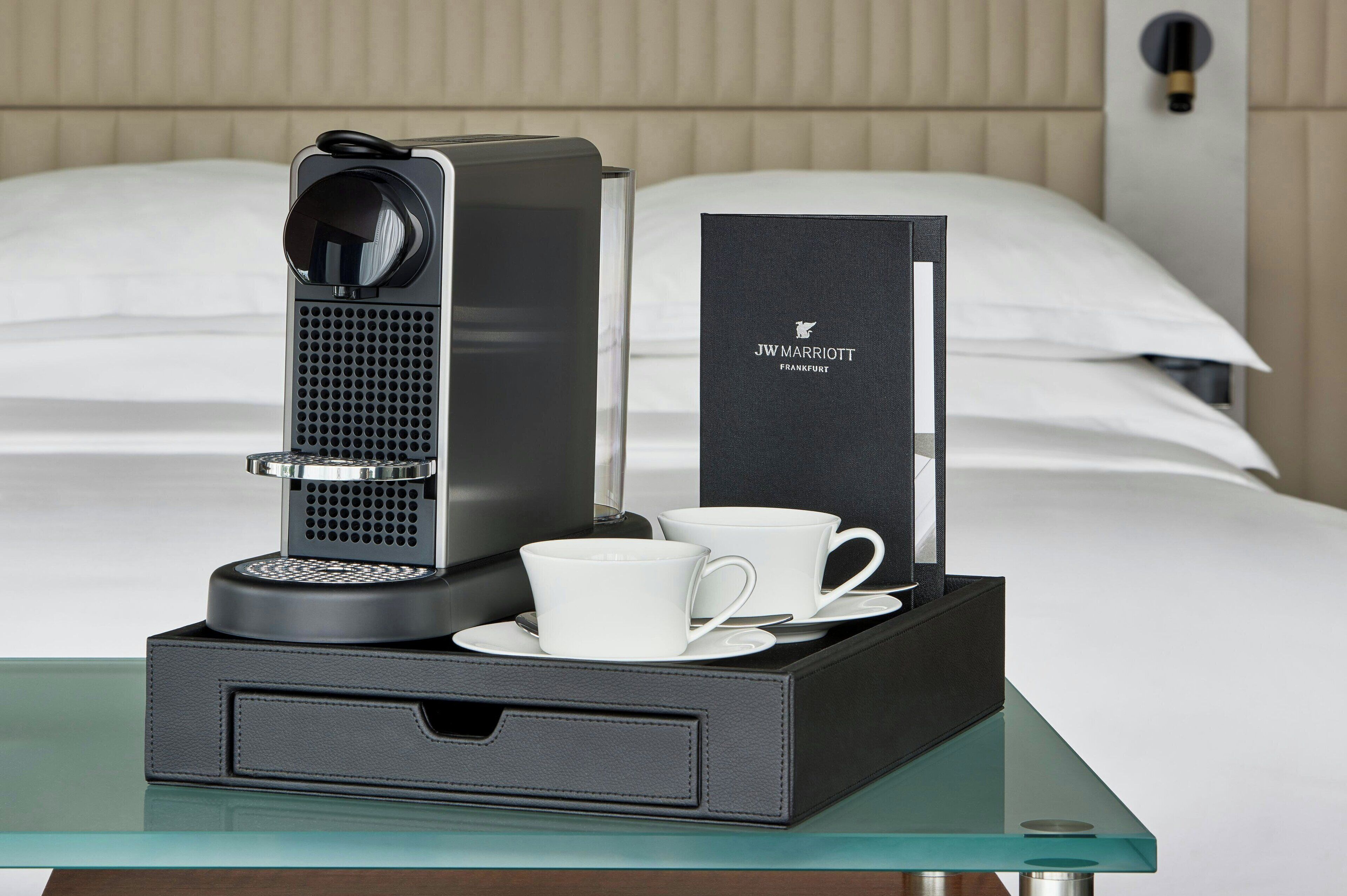 Habitación de hotel con cama, mesita de noche con cafetera, dos tazas blancas y una caja negra con el nombre del hotel.