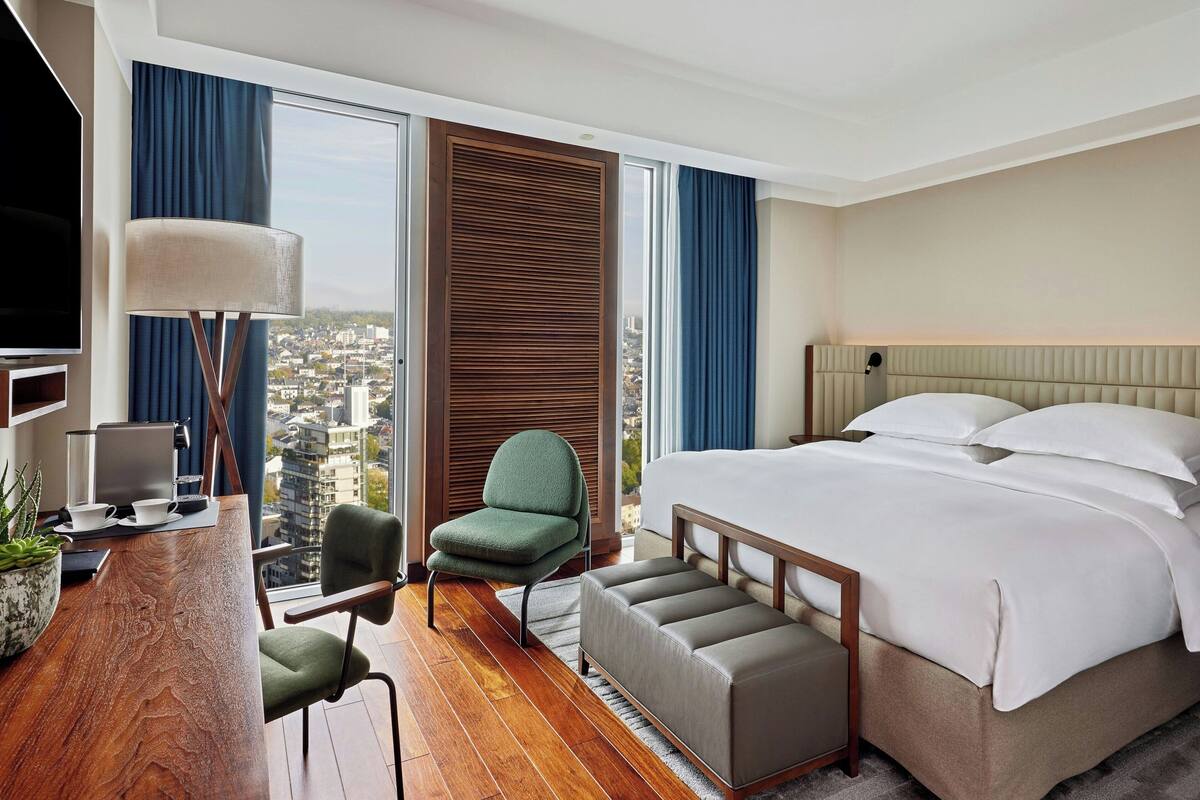 A hotel room with a large bed, a desk with a computer, a chair, and a view of the city through large windows.