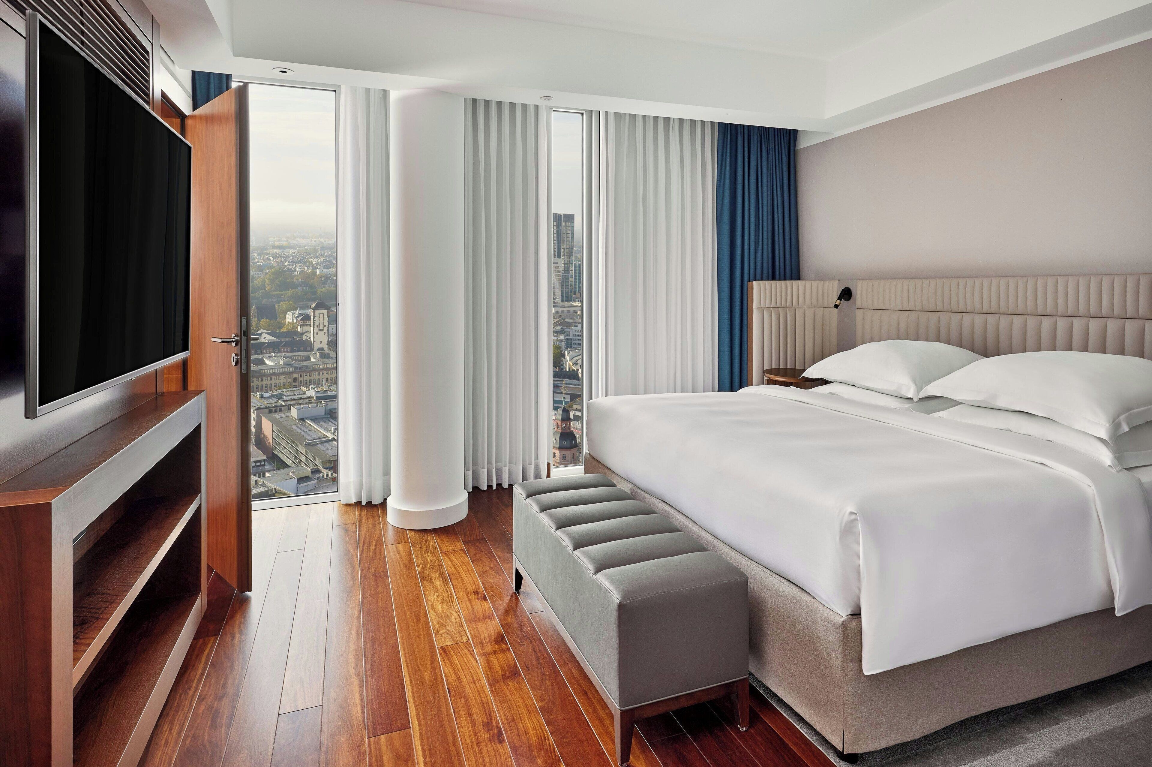 Quarto de hotel moderno com uma cama grande, televisão e vista da cidade através de janelas do chão ao teto.