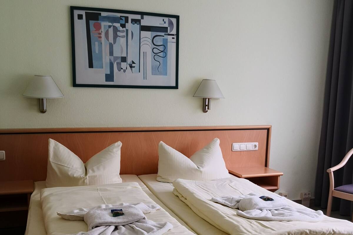 A hotel room with two beds, each with white linens and pillows, a wooden headboard, a framed abstract artwork above the beds, and two wall-mounted lamps.