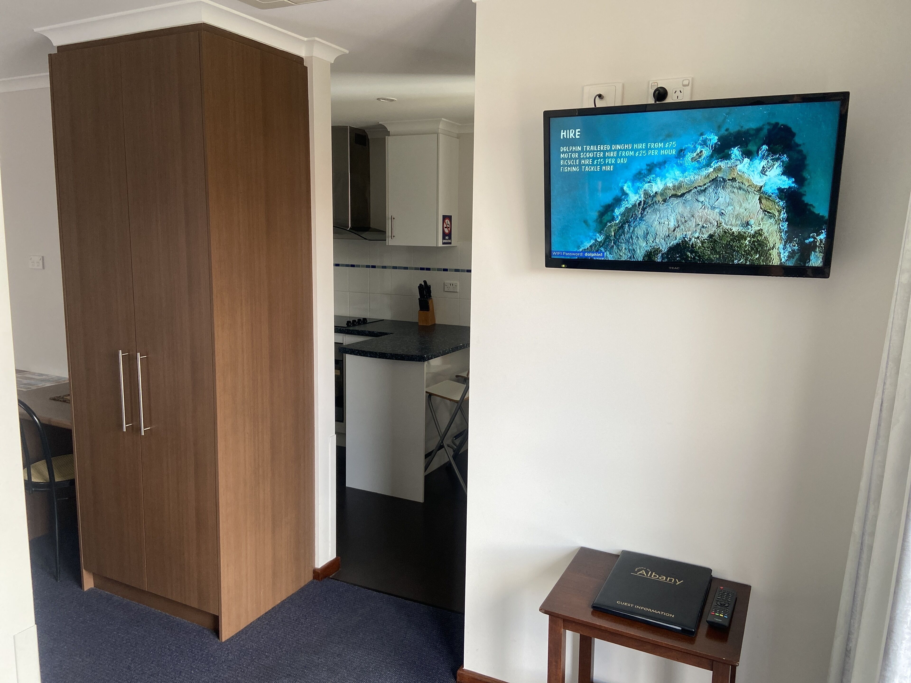Quarto com guarda-roupa de madeira, uma pequena mesa com um guia do hotel e uma televisão na parede exibindo serviços de aluguel de barco com golfinhos.