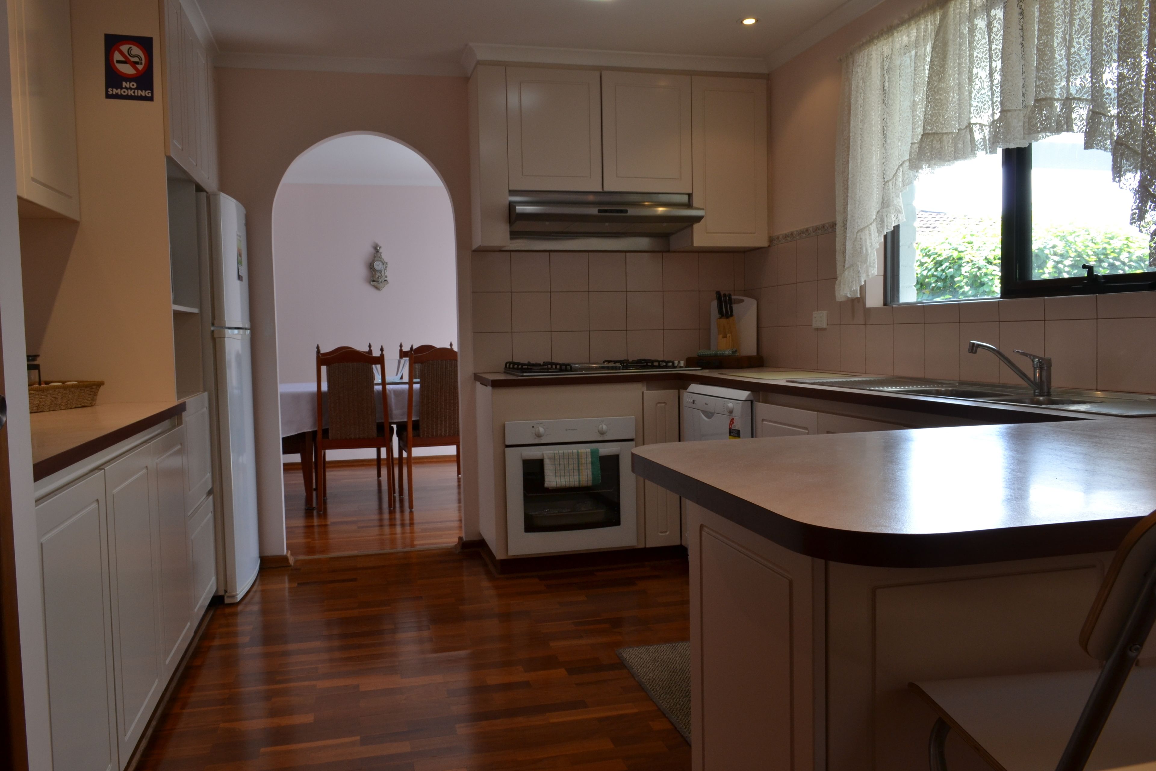 A kitchen with white cabinets, a stainless steel range hood, a window with lace curtains, and a wooden dining table with chairs.