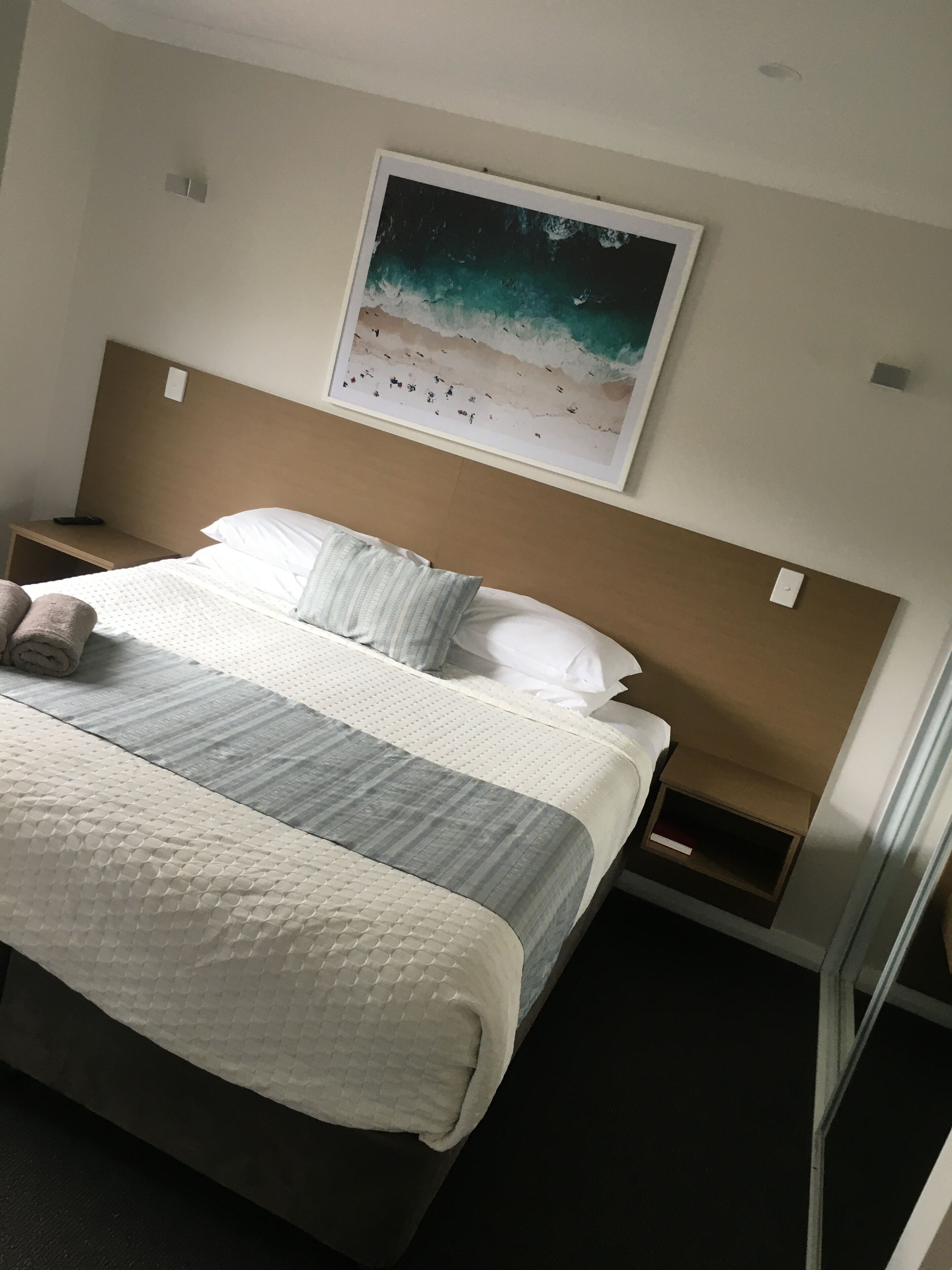 Et hotelværelse med en seng, sengegavl, et natbord, et indrammet billede af en strand og en lille hylde.