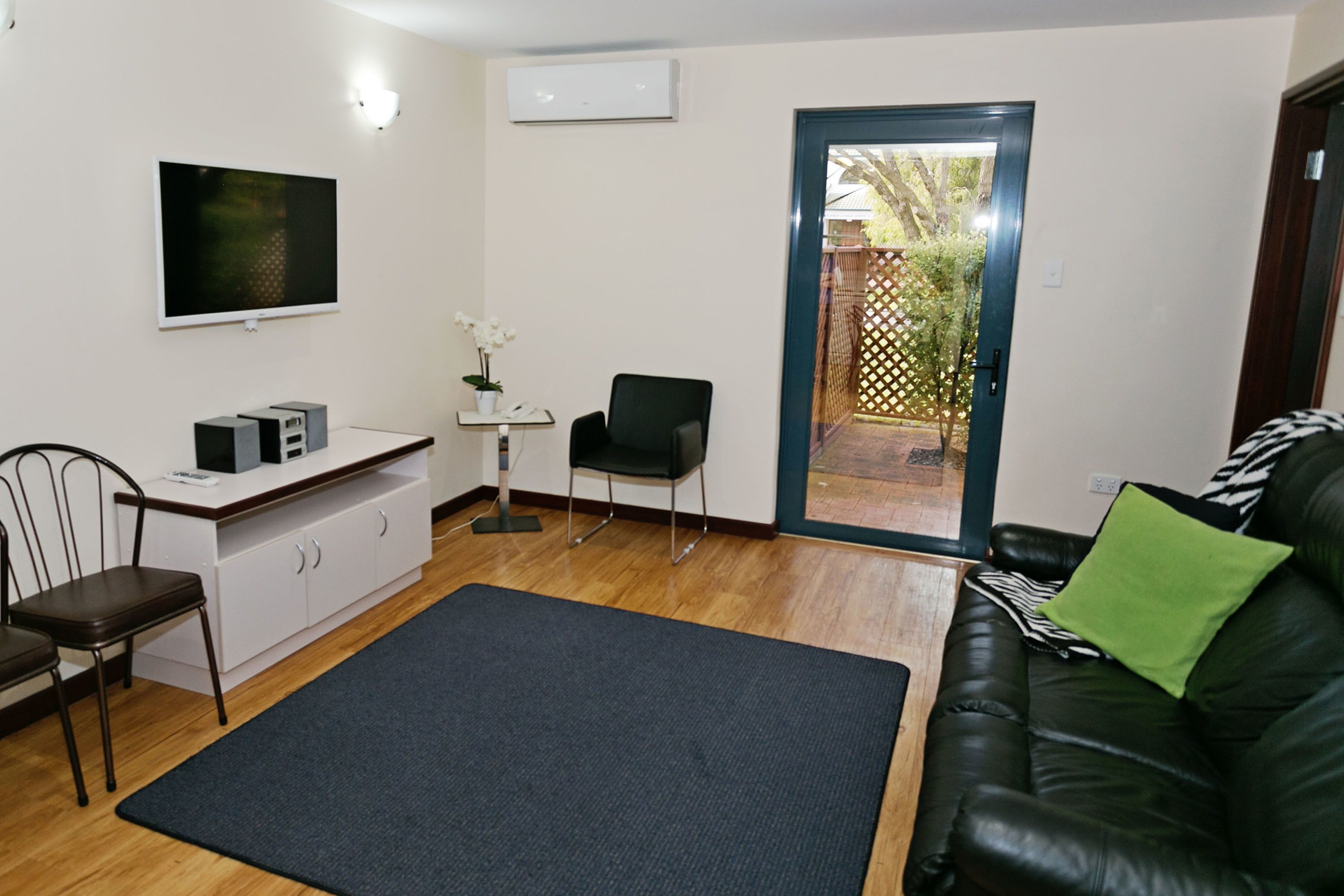 Ein modernes Wohnzimmer mit einer schwarzen Ledersofa, einem schwarzen Sessel, einem kleinen Schreibtisch mit Computer und Lautsprechern, einem flachbildschirm-Fernseher an der Wand und einem großen, dunklen Teppichboden.