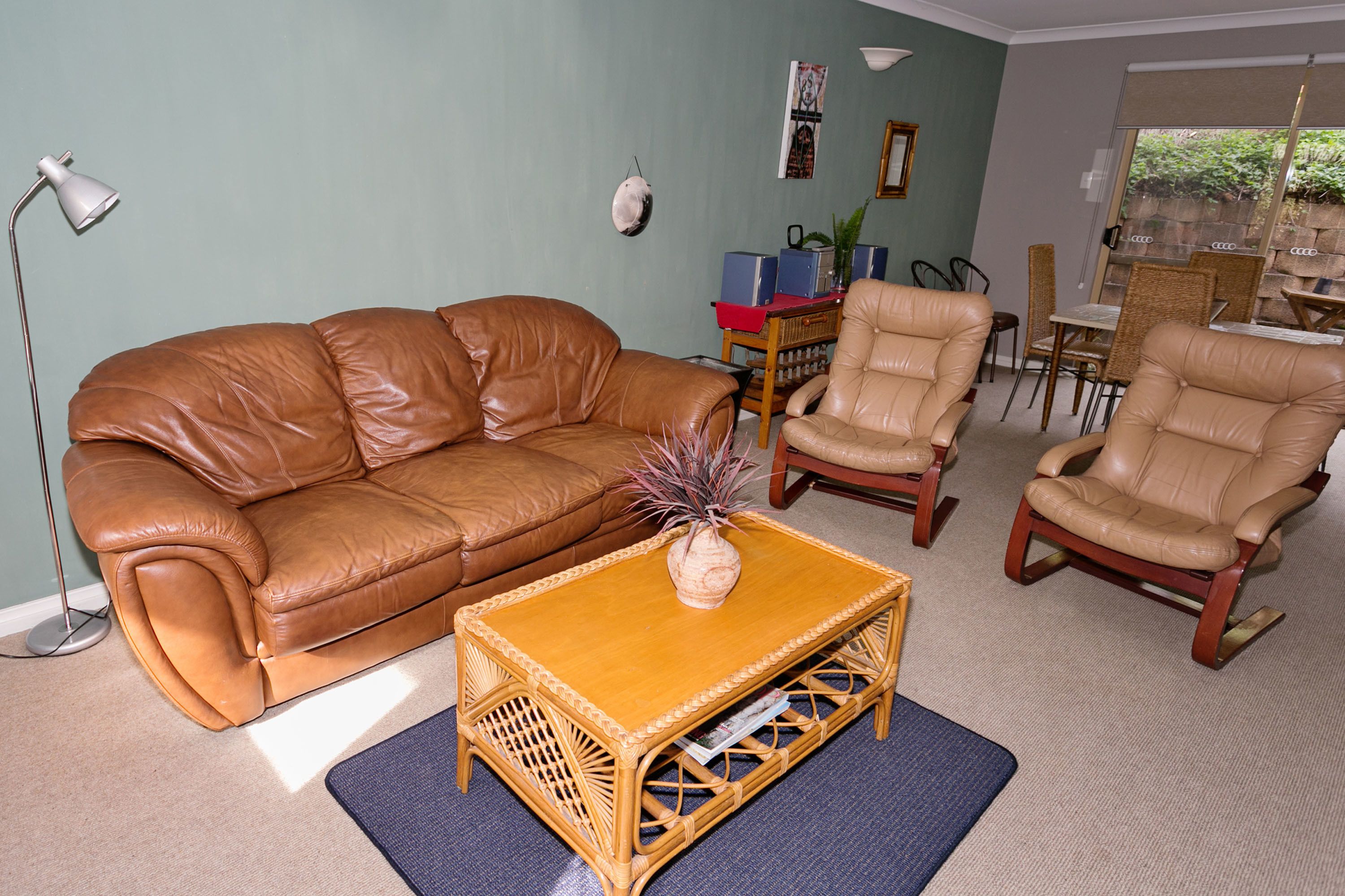 Un salón con un sofá de cuero marrón, dos sillones beige, una mesa de centro de madera con un florero y una lámpara de pie.