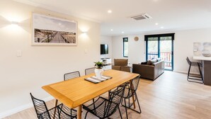 A modern living room with a wooden dining table, black wireframe chairs, a sofa, and a TV. - Dolphin Lodge (Middleton Beach)