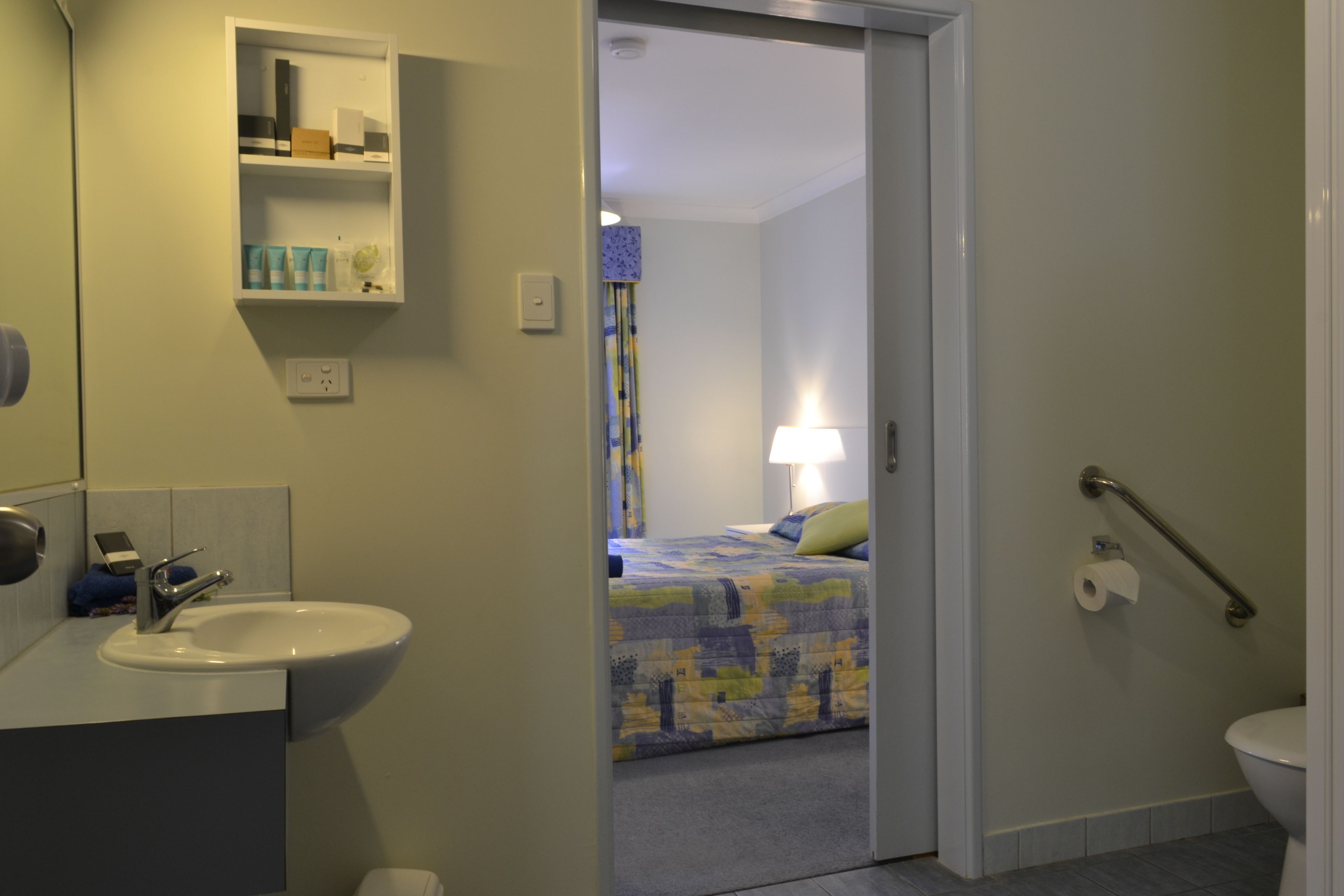 A bathroom with a sink, toilet, and a bed visible through an open door.