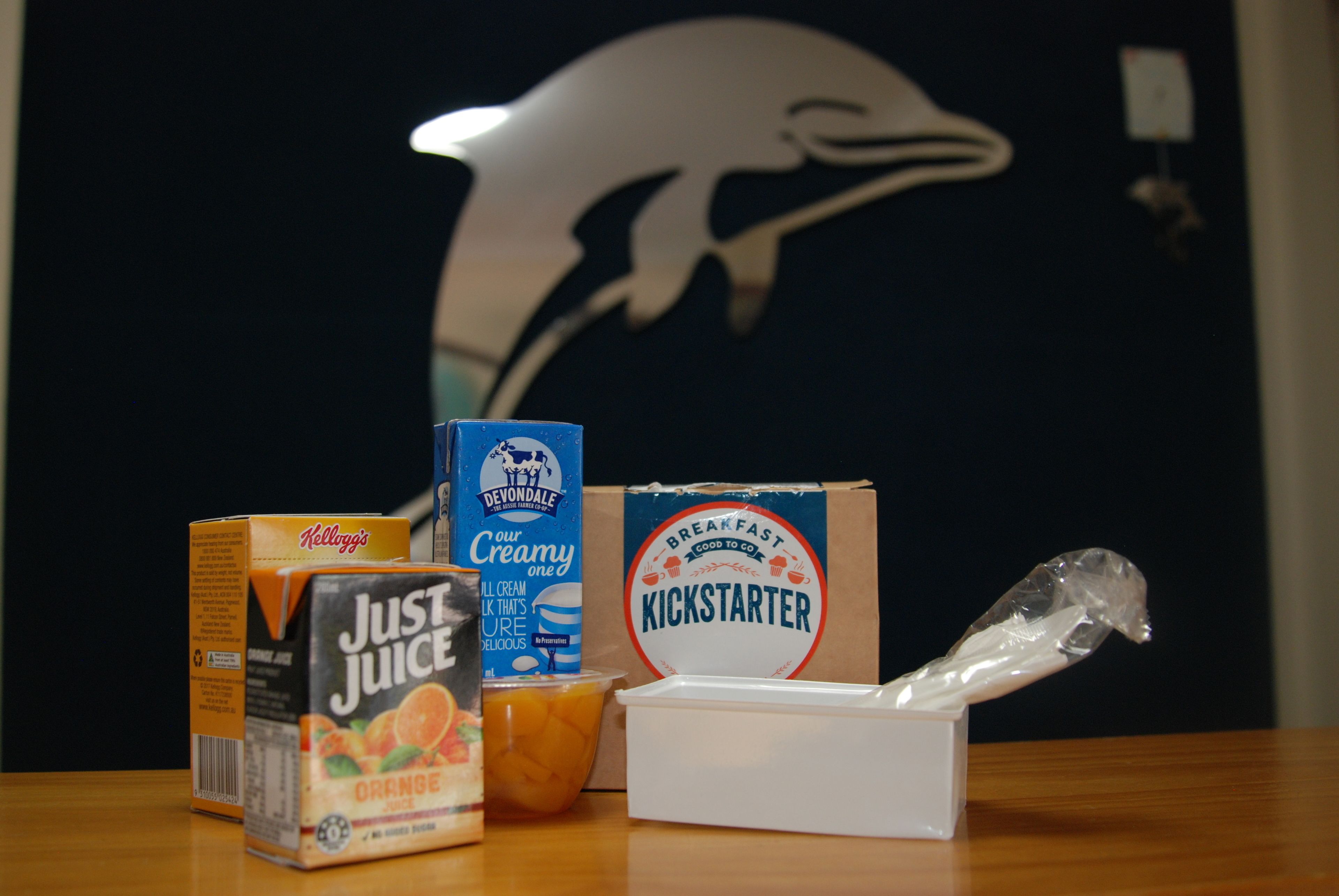 Una mesa con una caja de Just Juice, un cartón de Devondale's Creamy One, un envase de jugo de naranja, una caja de Kickstarter y una bolsa de plástico.
