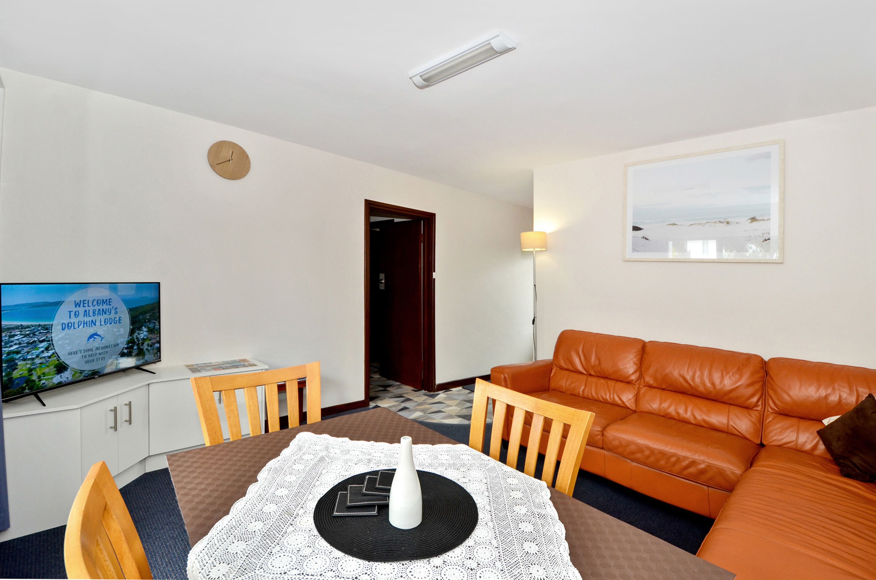 A living room with a brown leather sofa, a dining table with a white lace tablecloth, a flat-screen TV, and a clock on the wall.