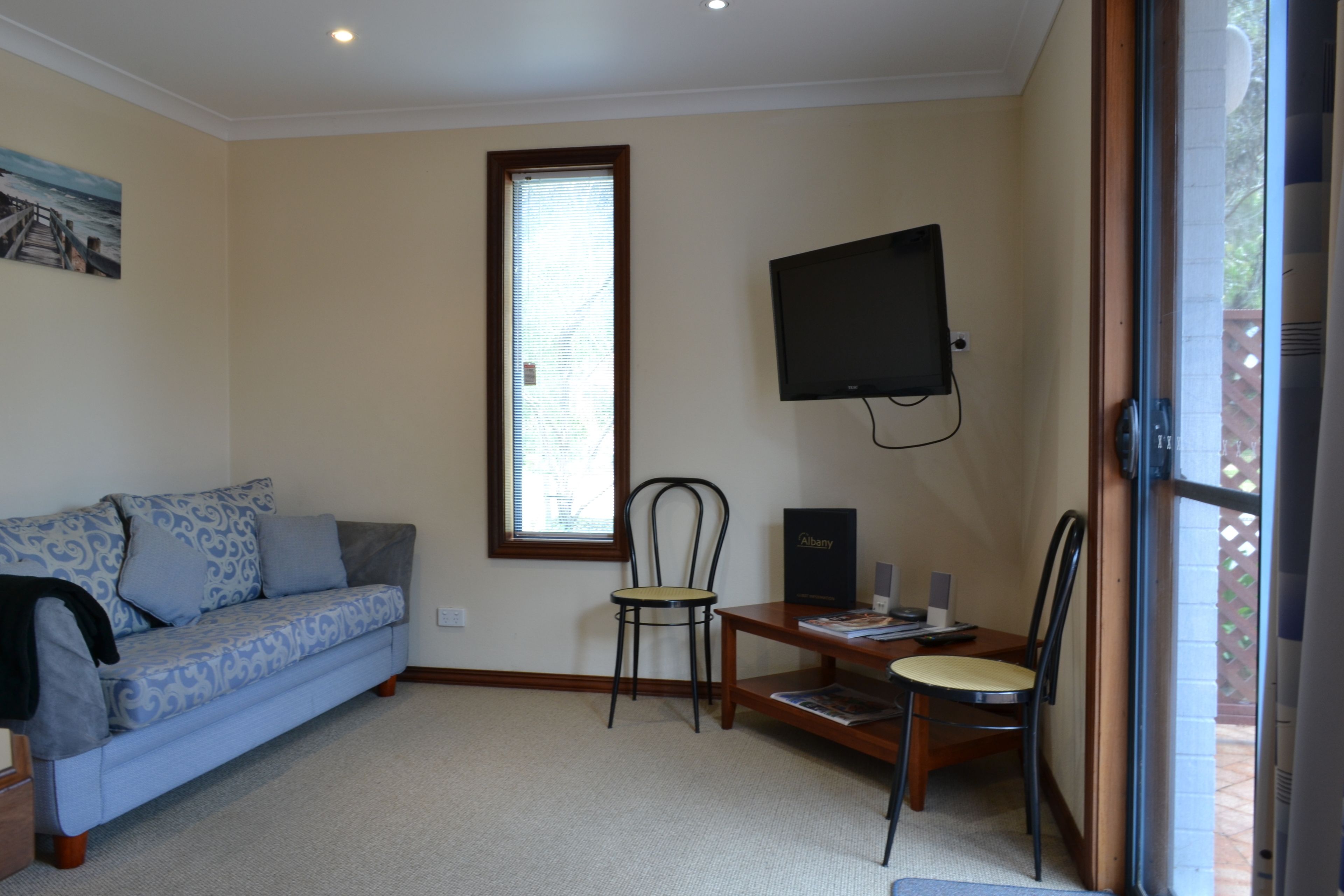 Una habitación con sofá, una silla, una mesita, televisión y ventana con persianas.