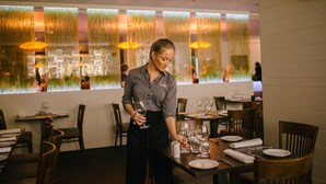 Een restaurant met een modern interieur, met houten tafels en stoelen, en een decoratief raam met verticale jaloezieën.