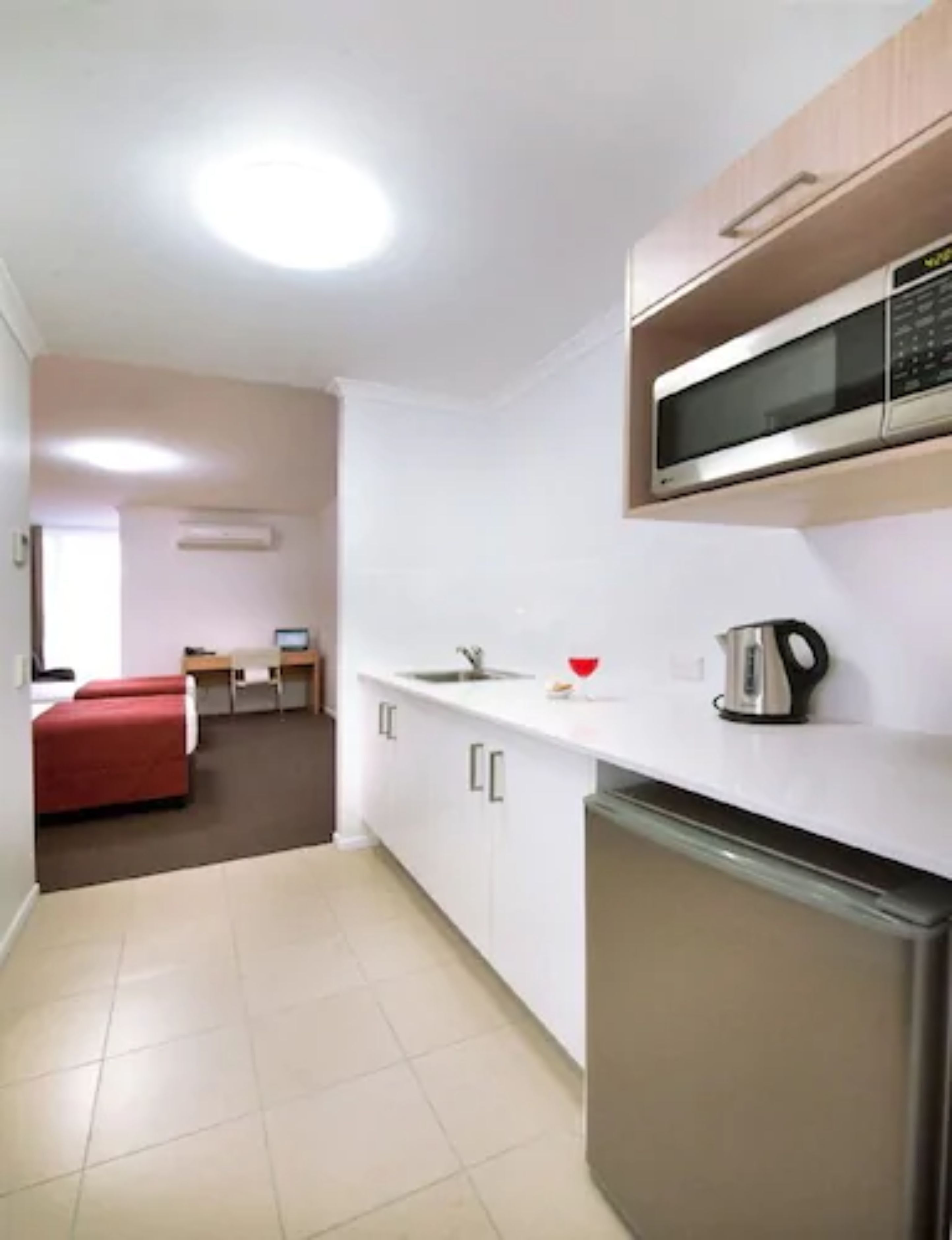 A compact kitchen with a microwave, sink, and a small refrigerator. There is a red couch and a desk in the adjacent room.
