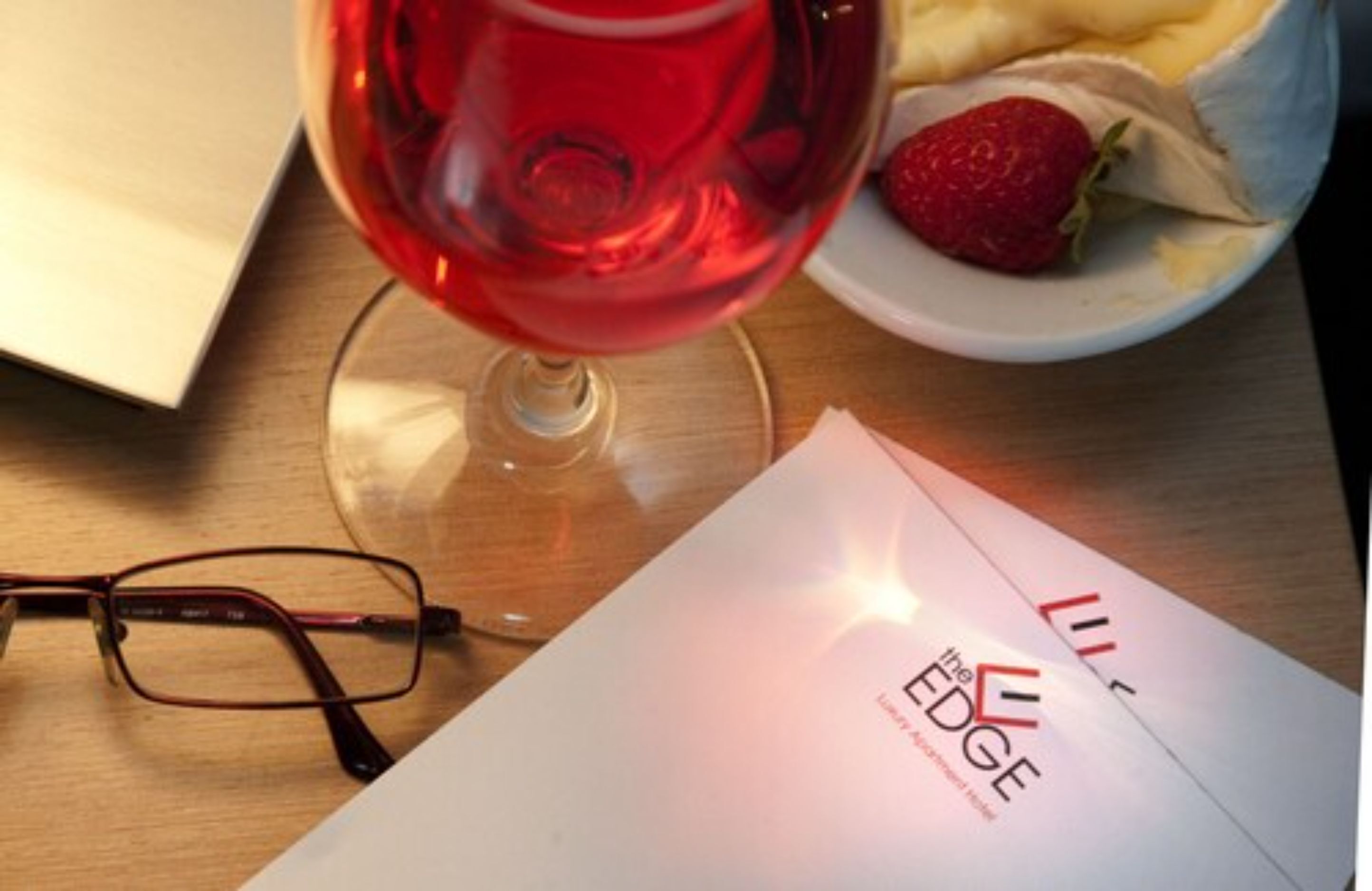 A glass of red wine, a pair of glasses, a plate with a strawberry, and a booklet titled "The EDGE".