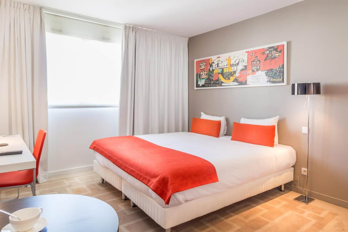 A hotel room with a bed, a desk, a chair, a table, and a floor lamp.