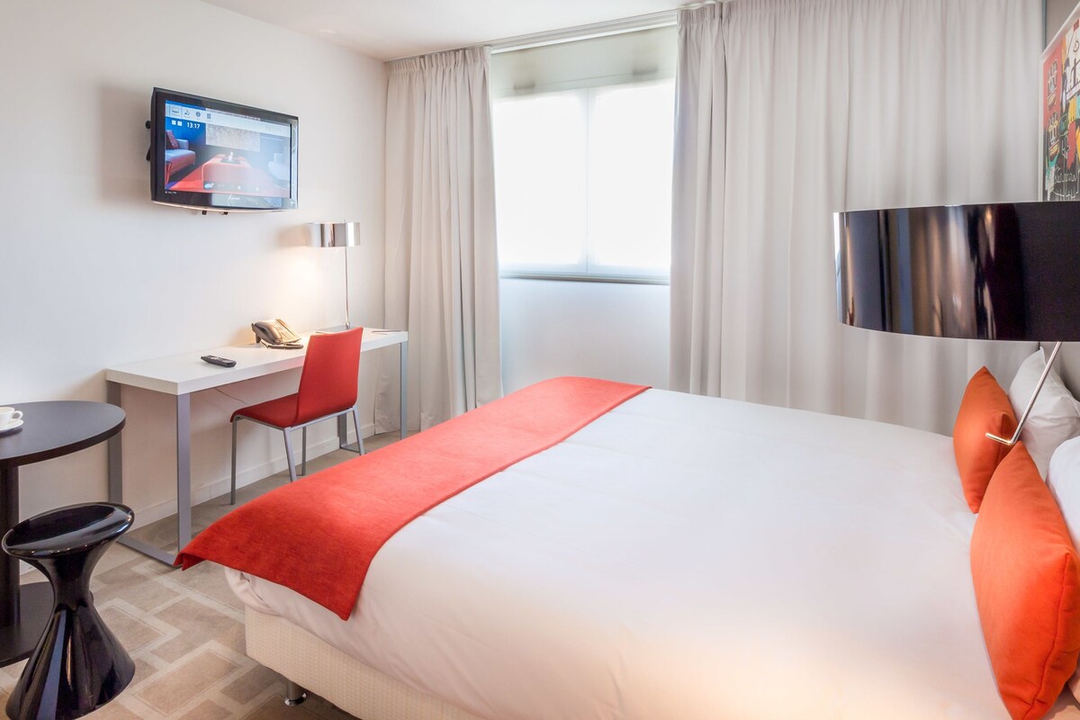 Habitación de hotel con cama, escritorio, silla, televisor y ventana con cortinas.