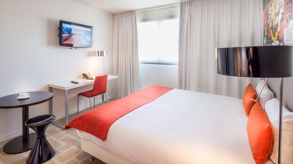 A hotel room with a bed, a desk, a chair, a TV, and a window with curtains.
