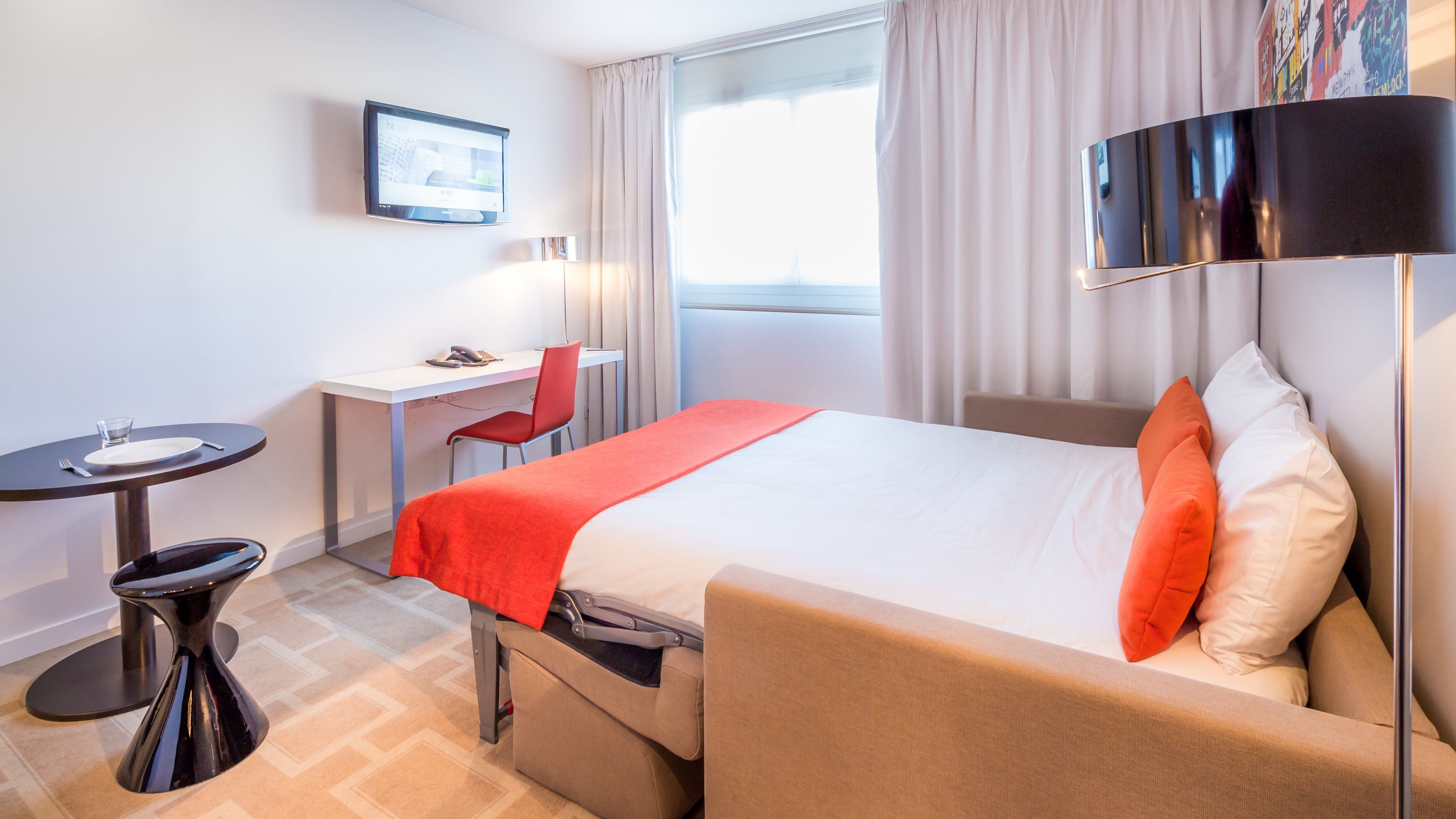 A hotel room with a bed, a desk, a chair, a small table, and a TV on the wall.