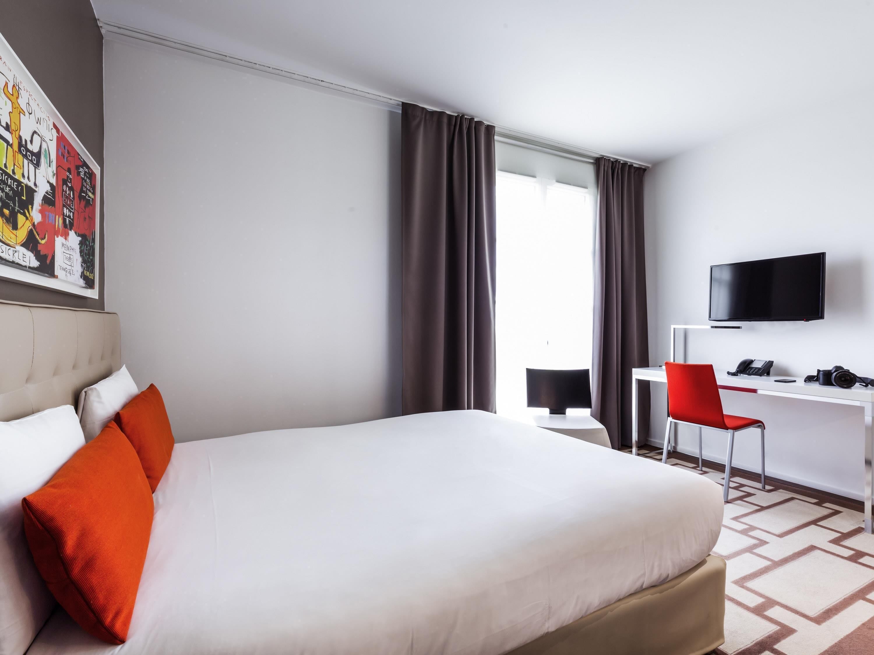 Ein Hotelzimmer mit Bett, Schreibtisch mit Stuhl, Fernseher und einem Fenster mit Vorhängen.