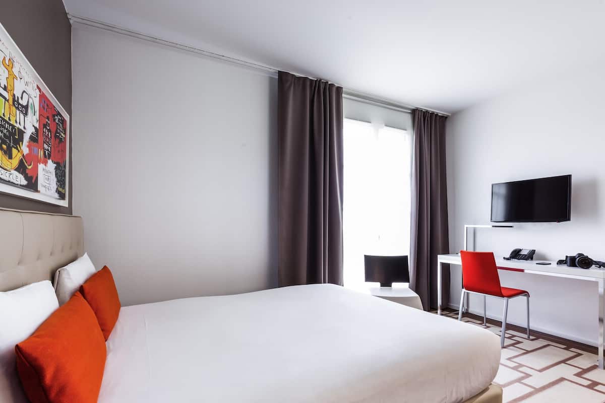 A hotel room with a bed, a desk with a chair, a television, and a window with curtains.