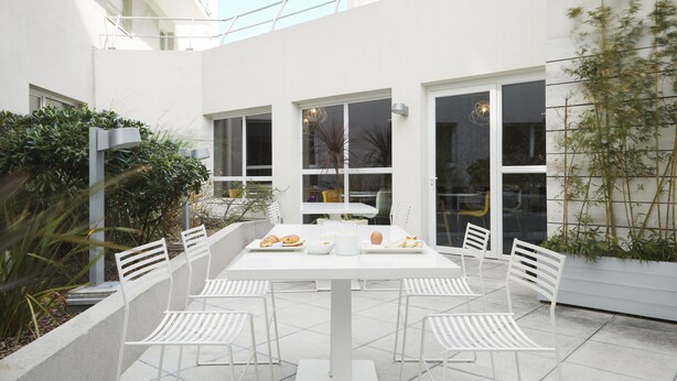 Un espacio moderno para comer al aire libre, con una mesa y sillas blancas, rodeado de grandes ventanales y follaje.
